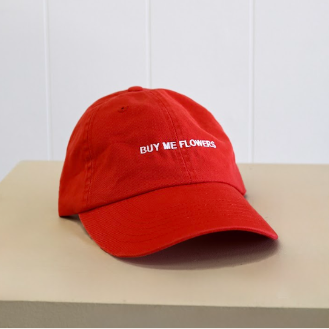 A red baseball cap with 'BUY ME FLOWERS' embroidered in white on the front, same-day delivery Perth