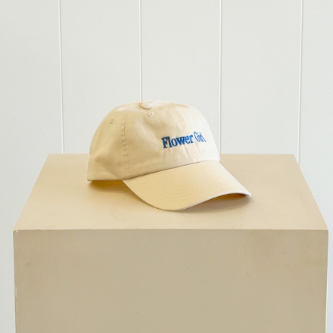 A beige baseball cap with blue embroidered text reading 'Flower Girl.' Same-day delivery Perth.
