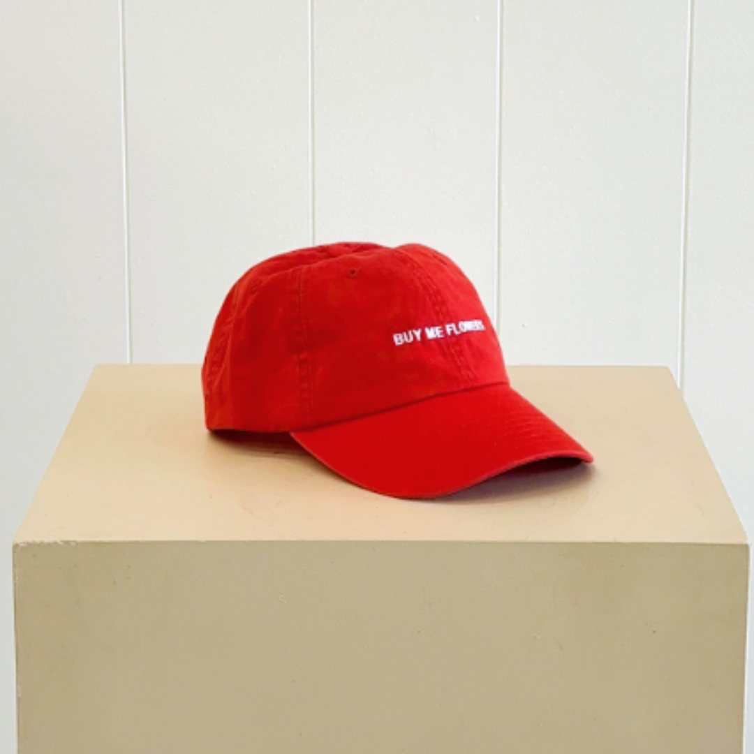 A red baseball cap with 'BUY ME FLOWERS' embroidered in white on the front, same-day delivery Perth