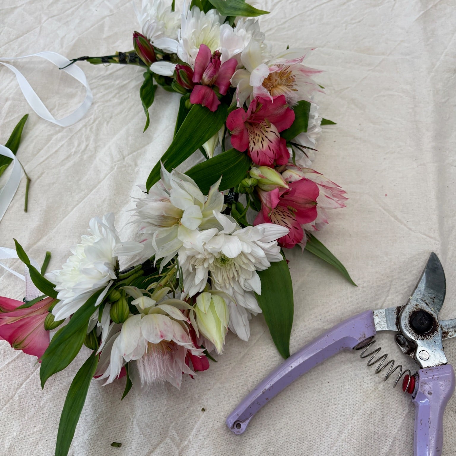 Floral arrangement with pink and white flowers and green leaves on a textured surface, accompanied by purple pruning shears.